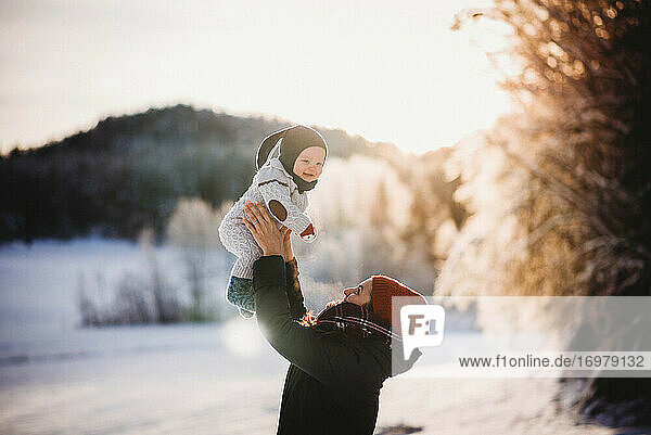 Mum holding smiling baby up in winter on sunny snowy day in the forest