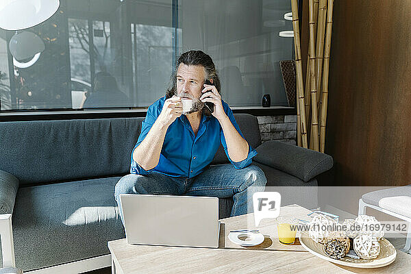 Mature man with beard talking on the phone and drinking coffee sitting on a sofa in front of his laptop looking out the window. Business concept