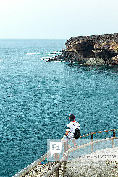 Junger Mann  der an einem Sommertag auf einer Klippe vor dem Meer sitzt.