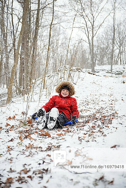 Happy boy sledding down a hill in the woods on a snowy winter day.