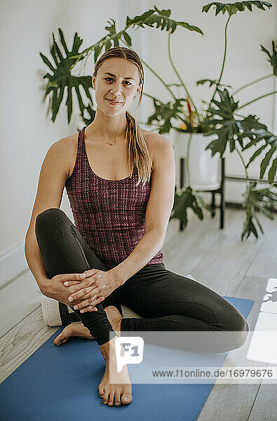 Porträt einer jungen  fitten weißen Frau auf einer Yogamatte sitzend im Haus