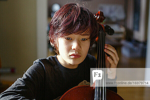 Ein Junge mit intensivem Fokus und rot gefärbtem Haar spielt Cello im Fensterlicht