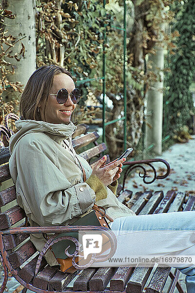 Entspannte junge Frau sitzt auf einer Bank und benutzt ein Mobiltelefon