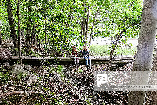 two girls sitting together and resting on bridge in woods while hiking