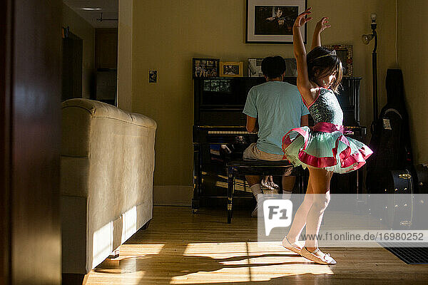 Ein kleines Mädchen im Tütü hebt den Arm und tanzt zur Klaviermusik des Vaters
