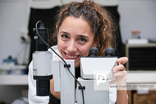 Junge Wissenschaftlerin schaut in die Kamera und lächelt neben einem Mikroskop in einem Labor. Laborforschung Konzept.