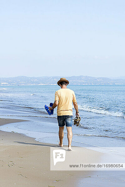 Rückansicht eines Mannes mit Hut und Schuhen  der barfuß am Meeresufer läuft