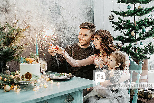 Junge Familie mit einer Tochter in festlicher Kleidung an einem gedeckten Tisch mit Kerzen  Girlanden  Wunderkerzen und einem Kuchen in der Nähe des Weihnachtsbaums an Silvester