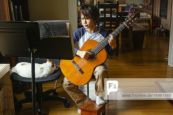 Ein Junge sitzt neben seiner Katze im Fensterlicht und übt zu Hause Gitarre