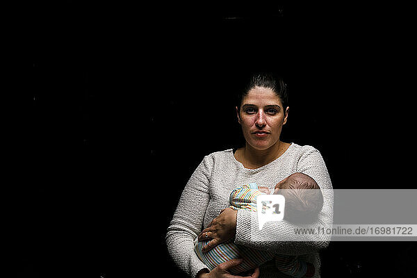 Ruhige Frau  die ein winziges neugeborenes Baby umarmt und in die Kamera schaut  während sie vor einem schwarzen Hintergrund steht