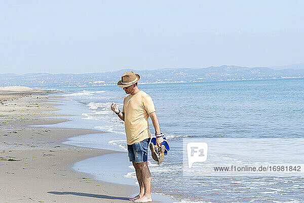 Mann steht am Meeresufer  hält Schuhe und betrachtet eine Muschel