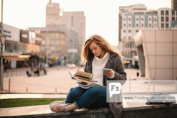 Serious teenage student girl learning while sitting in city in autumn