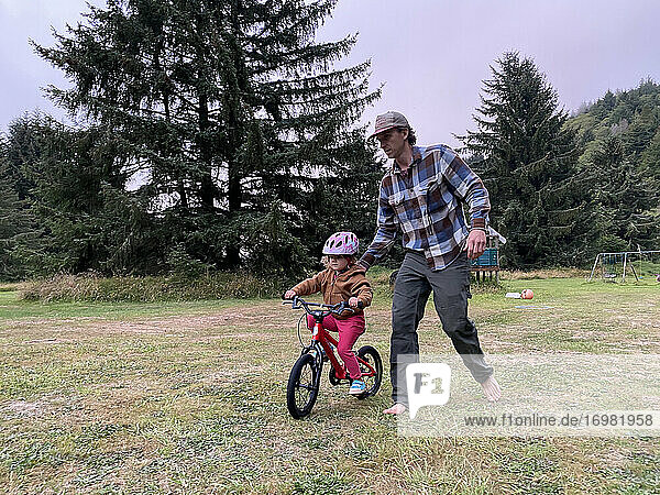 A father teaches his daughter how to ride a bike on a grass field.