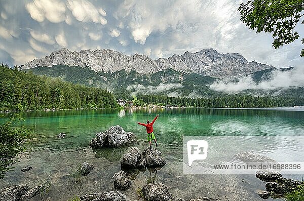 Young man sticks his arms in the air and stands on a rock on the shore  Eibsee lake in front of Zugspitze massif with Zugspitze  dramatic Mammaten clouds  Wetterstein range  near Grainau  Upper Bavaria  Bavaria  Germany  Europe