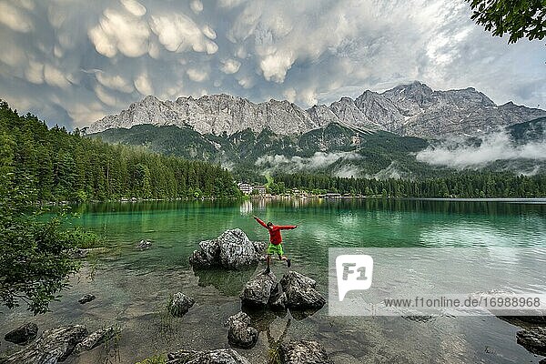 Young man jumps from rock to rock  Eibsee lake in front of Zugspitze massif with Zugspitze  dramatic Mammaten clouds  Wetterstein range  near Grainau  Upper Bavaria  Bavaria  Germany  Europe