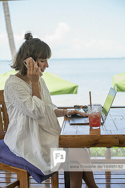 Frau mit Smartphone und Laptop arbeiten am Strand Cafe Tisch