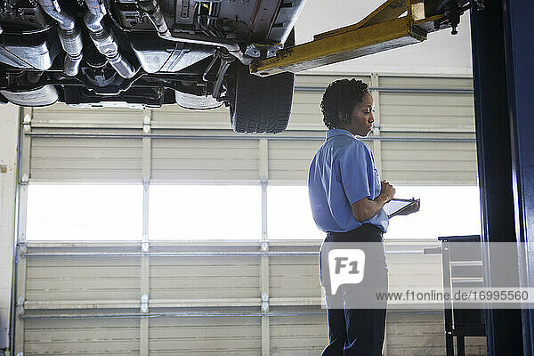 Weiblicher Mechaniker  der ein digitales Tablet hält  das unter einem Autochassis in einer Autowerkstatt steht