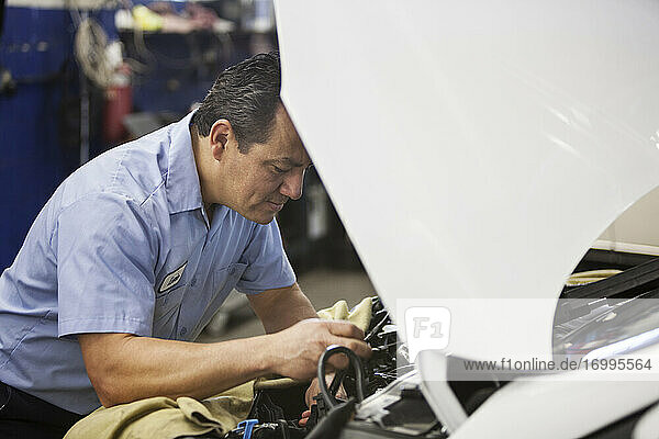 Ein hispanischer Mechaniker lehnt sich an ein Auto und arbeitet am Motorraum in einer Autowerkstatt