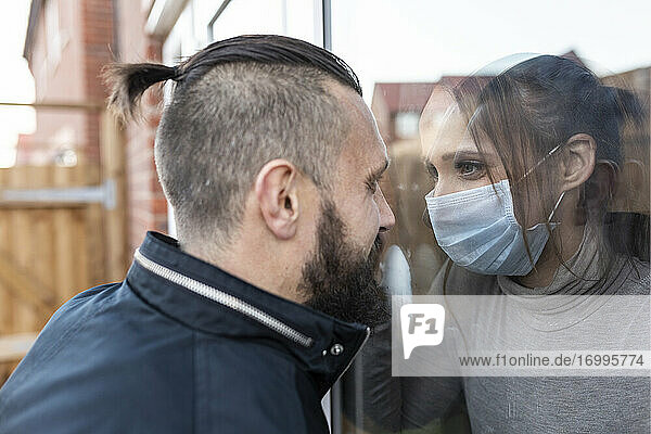 Freund betrachtet Freundin mit Gesichtsschutzmaske durch Fensterscheibe bei COVID-19