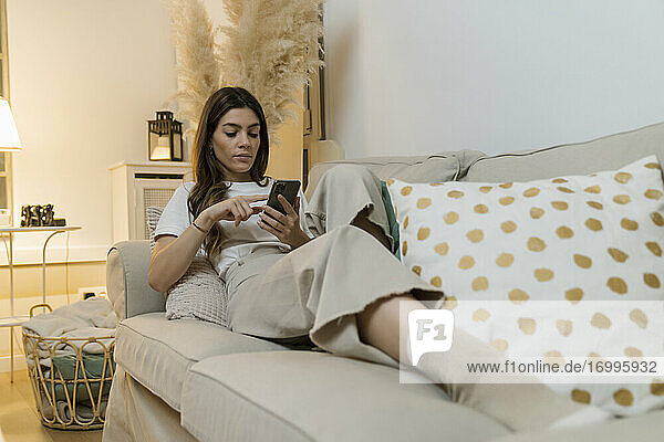 Junge Frau benutzt ein Mobiltelefon  während sie sich zu Hause auf dem Sofa entspannt