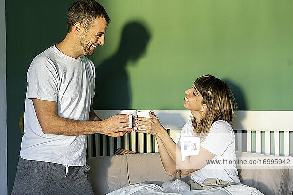 Lächelnder Freund und Freundin stoßen mit Kaffee an  während sie zu Hause auf dem Bett sitzen