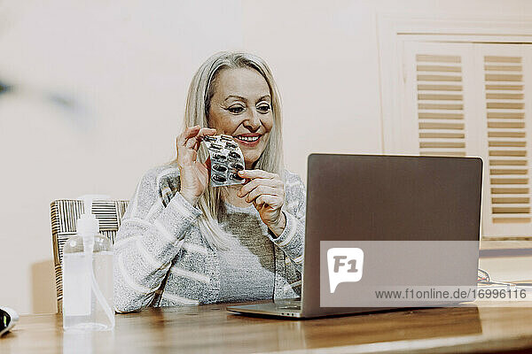 Lächelnde ältere Frau  die während einer Online-Konsultation zu Hause Medikamente zeigt