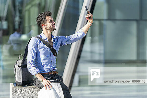 Lächelnder Geschäftsmann  der ein Selfie mit seinem Mobiltelefon macht  während er im Freien sitzt