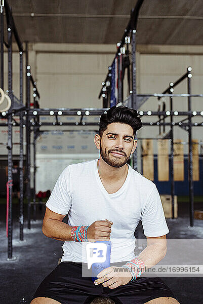 Lächelnder Mann  der eine Wasserflasche hält  während er im Fitnessstudio sitzt
