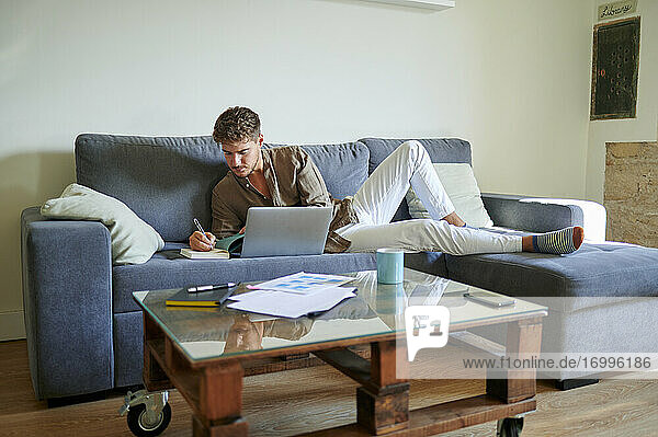 Junger männlicher Freiberufler  der im Wohnzimmer auf dem Sofa liegend mit seinem Laptop in sein Tagebuch schreibt