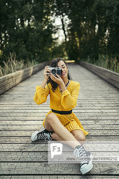 Junge Fotografin in gelbem Kleid und schwarzen Turnschuhen hält eine analoge Kamera auf einer Holzpromenade