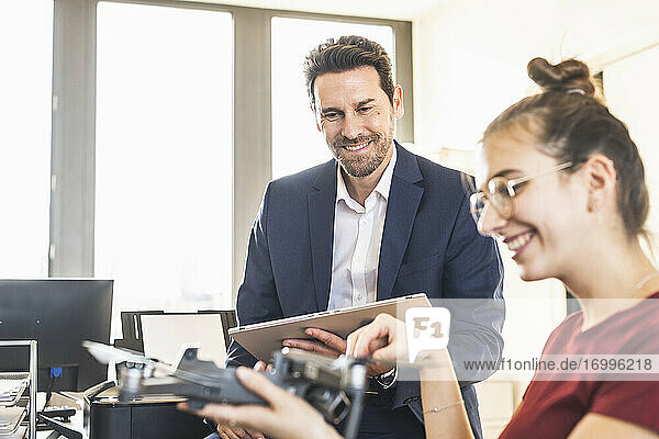Lächelnder Geschäftsmann mit digitalem Tablet und Blick auf einen Kollegen  der einen Quadcopter hält  während er im Büro sitzt