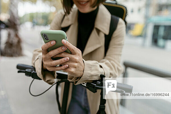 Frau  die ein Smartphone benutzt  während sie mit einem elektrischen Tretroller auf der Straße steht