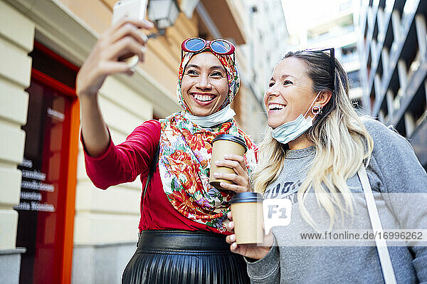 Female friends taking selfie while holding coffee cups in city during COVID-19