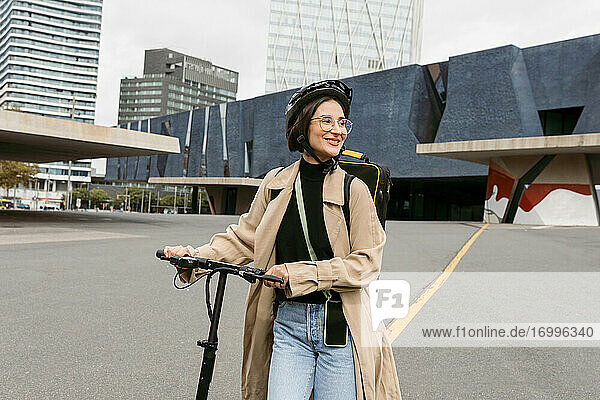 Lächelnde Frau mit Fahrradhelm  die wegschaut  während sie mit einem Elektroroller in der Stadt spazieren geht