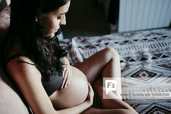Schwangere Frau berührt den Bauch  während sie zu Hause auf dem Bett sitzt