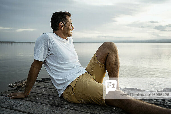 Älterer Mann verbringt seine Freizeit am Steg am See gegen den Himmel sitzend
