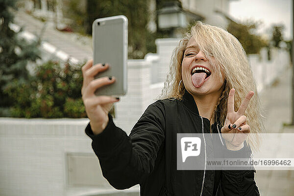 Glückliche junge Frau  die ein Selfie macht  während sie friedlich gestikuliert und die Zunge durch ihr Smartphone herausstreckt