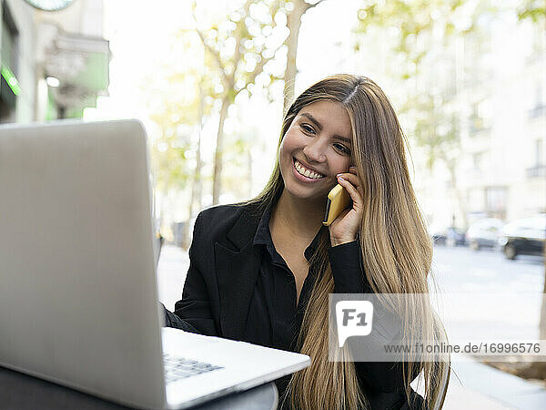 Junge Frau  die in einem Café am Straßenrand sitzt und mit ihrem Laptop telefoniert