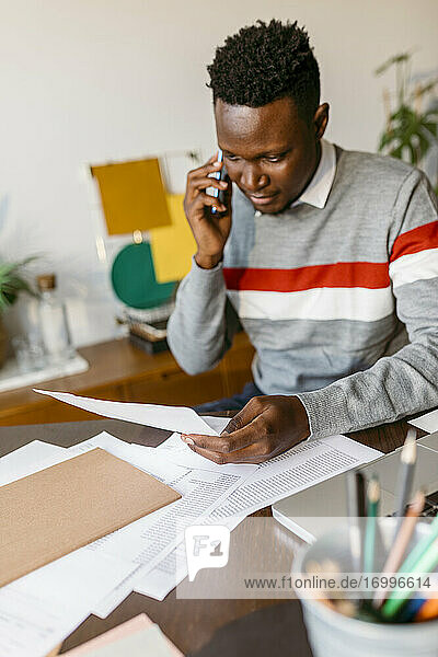Male entrepreneur talking on smart phone while holding document in home office