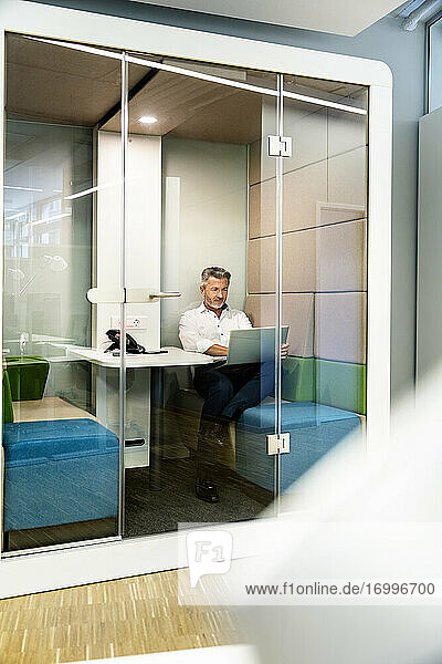 Männlicher Unternehmer arbeitet am Laptop  während er in einer Kabine im Büro sitzt