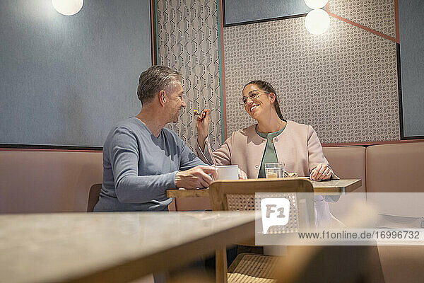 Smiling businesswoman feeding man while sitting at modern cafe