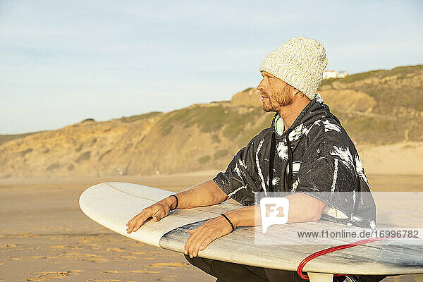 Mann schaut weg  während er mit einem Surfbrett am Strand hockt
