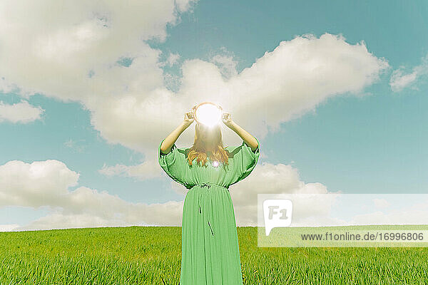 Junge Frau in grünem Kleid steht auf einem Feld und hält einen reflektierenden Spiegel
