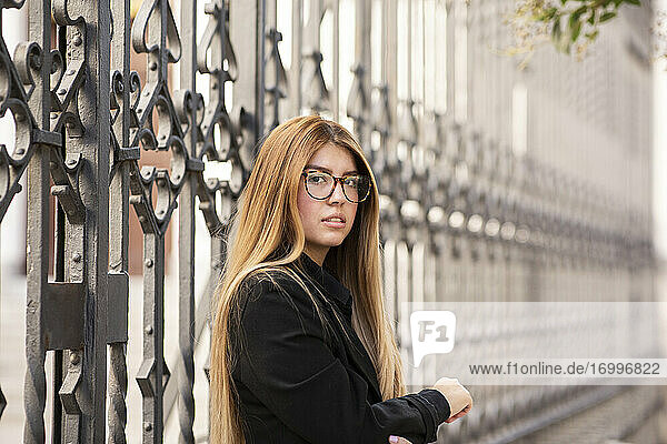 Young woman wearing eyeglasses standing against gate