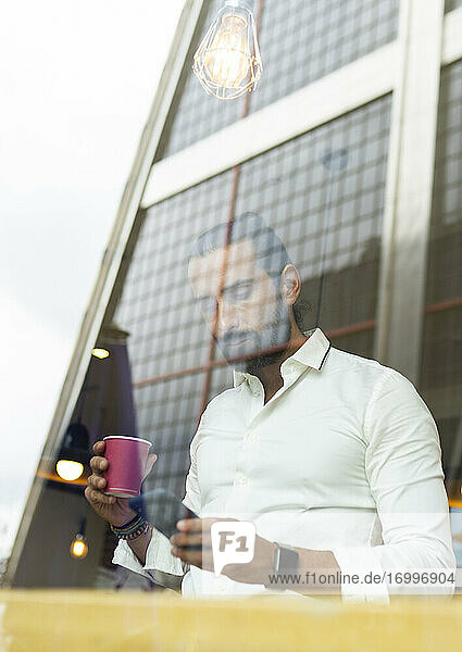 Candid portrait of bearded businessman drinking coffee and using phone inside cafe