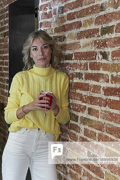 Fashionable mature woman holding tea cup while leaning on brick wall in kitchen
