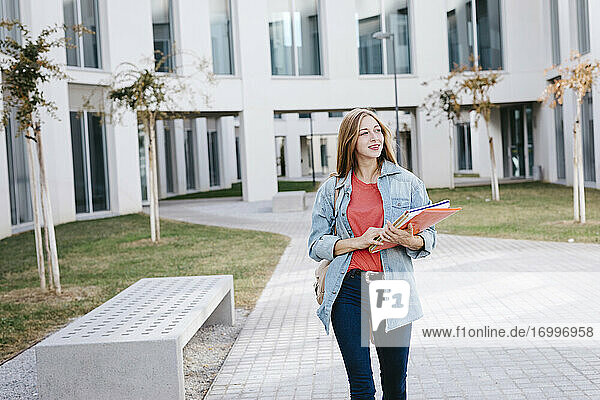 Lächelnde schöne blonde Studentin  die wegschaut  während sie auf dem Universitätscampus spazieren geht