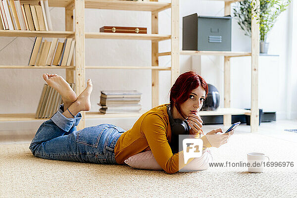 Rothaarige Frau  die ein Mobiltelefon benutzt  während sie zu Hause auf dem Teppich liegt