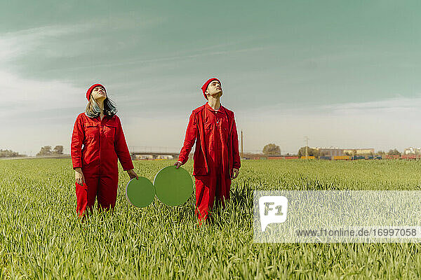 Junges Paar in roten Overalls steht auf einem Feld mit zwei grünen Kreisen und schaut nach oben