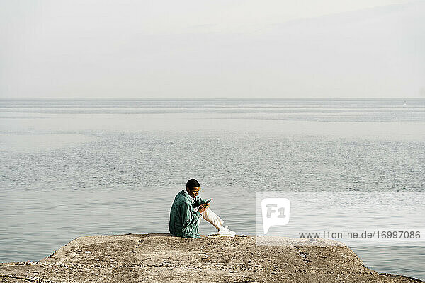 Junger Mann  der ein Mobiltelefon benutzt  während er auf einem Pier am Meer und Himmel sitzt